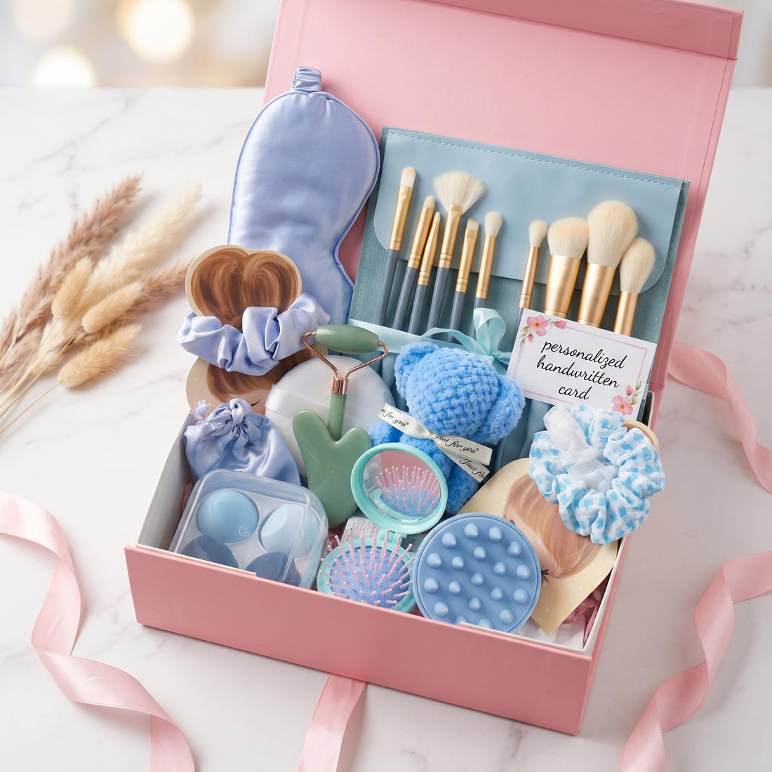 May include: A pink gift box containing a light blue sleep mask, a set of makeup brushes in a blue case, a green jade roller, a blue crochet bear, a blue hair scrunchie, a blue patterned scrunchie, a small mirror, a scalp massager, and a card.