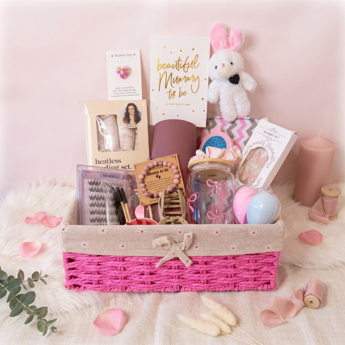 May include: A gift basket with a bright pink woven base and beige fabric trim containing a heatless curling set, a white plush bunny, a glass tumbler with pink bows, a Mama to be beaded bracelet, a card, false eyelashes, and beauty accessories.