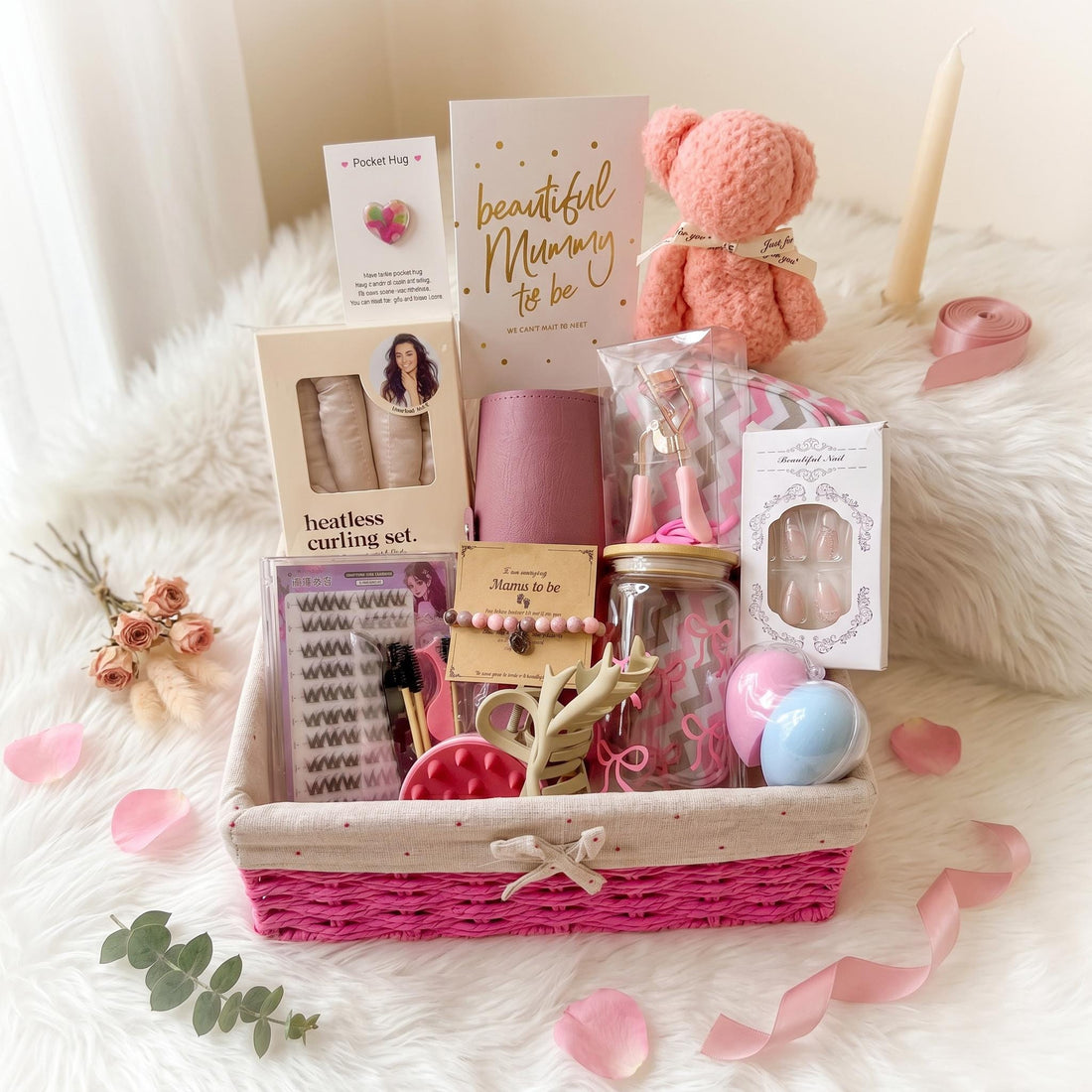 May include: A gift basket with a bright pink woven base and beige fabric trim containing a heatless curling set, a white plush bunny, a glass tumbler with pink bows, a Mama to be beaded bracelet, a card, false eyelashes, and beauty accessories.